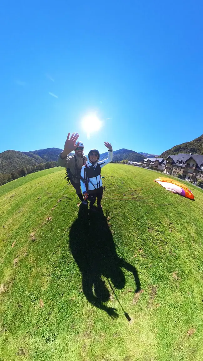 Piloto y pasajera saludando sobre la hierba junto al parapente, foto de Aran Parapent para Vuelo Luchon