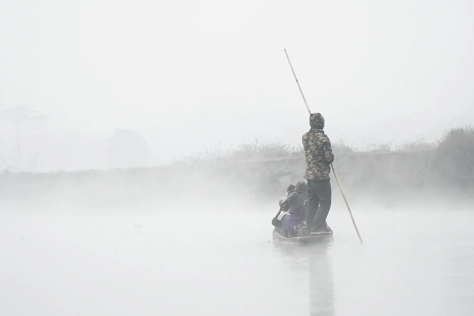 Un barquero guía una canoa de madera con pértiga entre la niebla del río, con dos pasajeros